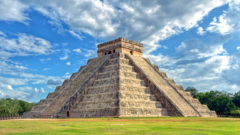 Famous-mayan-step-pyramid-of-El-Castillo-stands-in-grassy-field-with-a-cloudy-blue-sky