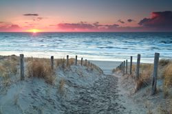 A wooden fenced lined path through sand dunes leads to a peaceful beach with small waves at sunset