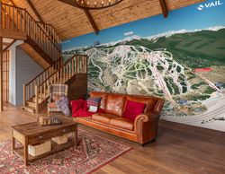 A-mural-of-the-Vail-mountain-ski-trail-map-in-a-cabin-style-living-room-with-a-leather-couch-plaid-wingback-chair-wood-coffee-table-on-an-ornate-rug-wood-ceiling-floors-and-staircase