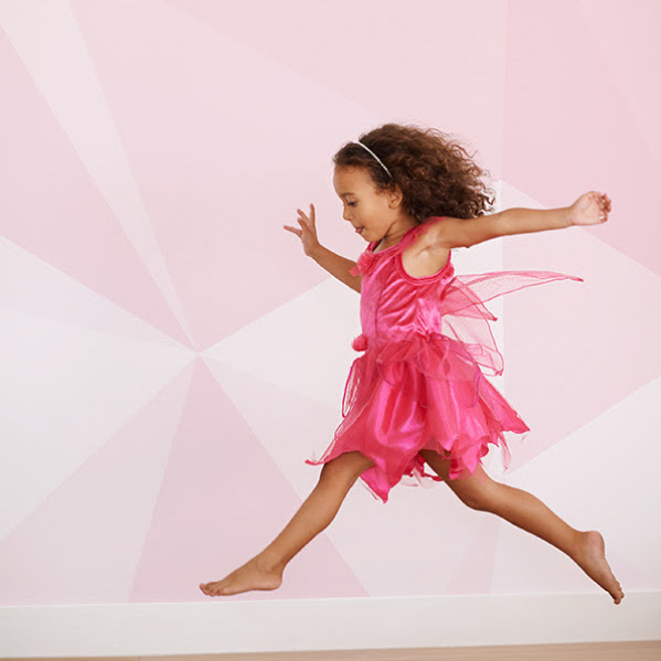 Little girl in a pink dress jumping in front of a wall with pink geometric wallpaper