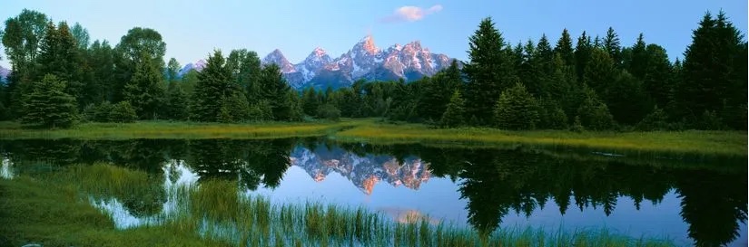 Panoramic view of the peaks of Grand Teton peaks framed by forest reflected in a lake