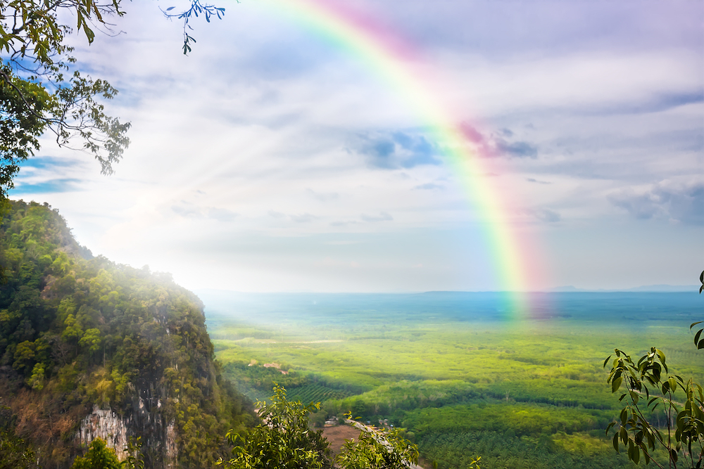 Sun bursts from behind a densely forested mountain creating a rainbow over the valley