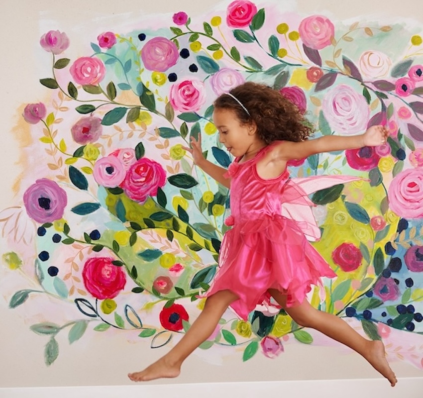 A little girl in a pink dress is jumping in front of a floral wall mural.